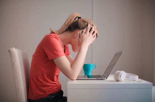 Teenage girl with blonde hair sits at a desk with her head in her hands. There is a laptop, a blue mug and a pair of headphones on the desk.